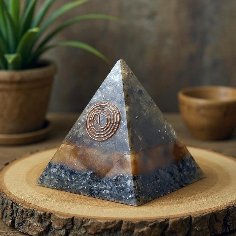 May include: A layered pyramid-shaped decorative object with a copper spiral design. The pyramid features a combination of gray, brown, and blue tones. It is displayed on a wooden slice, with a plant and a wooden bowl in the background.