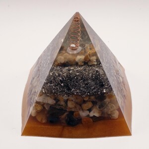 Puede incluir: Una escultura piramidal de resina con una espiral de cobre en la parte superior. La pir&aacute;mide est&aacute; rellena de capas de peque&ntilde;as piedras y material oscuro y granulado. La base es de color &aacute;mbar c&aacute;lido, creando una pieza decorativa &uacute;nica.
