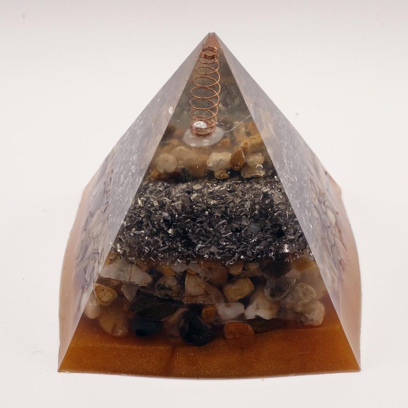 Puede incluir: Una escultura piramidal de resina con una espiral de cobre en la parte superior. La pir&aacute;mide est&aacute; rellena de capas de peque&ntilde;as piedras y material oscuro y granulado. La base es de color &aacute;mbar c&aacute;lido, creando una pieza decorativa &uacute;nica.