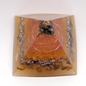 Peut inclure: Une pyramide &agrave; base carr&eacute;e avec un ext&eacute;rieur en r&eacute;sine ambr&eacute;e transparente. &Agrave; l'int&eacute;rieur, des couches de fil de cuivre, des copeaux de m&eacute;tal et un amas d'objets m&eacute;talliques sombres au sommet. La pyramide a un design g&eacute;om&eacute;trique.