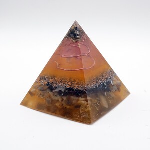 Peut inclure: Un objet d&eacute;coratif en forme de pyramide avec un ext&eacute;rieur en r&eacute;sine ambr&eacute;e transparente. L'int&eacute;rieur pr&eacute;sente des couches de pierres brunes, noires et blanches, avec un motif de fil rouge. La pyramide est sur fond blanc.