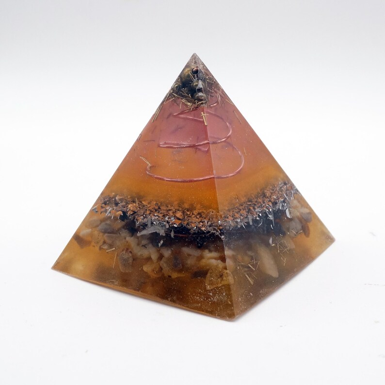 Peut inclure: Un objet d&eacute;coratif en forme de pyramide avec un ext&eacute;rieur en r&eacute;sine ambr&eacute;e transparente. L'int&eacute;rieur pr&eacute;sente des couches de pierres brunes, noires et blanches, avec un motif de fil rouge. La pyramide est sur fond blanc.