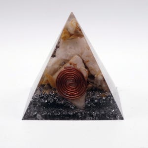 May include: A clear pyramid-shaped orgone energy generator. The pyramid contains white and yellow crystals, dark gray minerals, and a copper coil. The pyramid is on a white background.