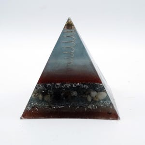 May include: A pyramid-shaped decorative object with a layered design. The top section is a gradient of blue and clear resin, followed by a red-brown layer, and a base with stones and dark inclusions. A coiled wire sits at the apex.