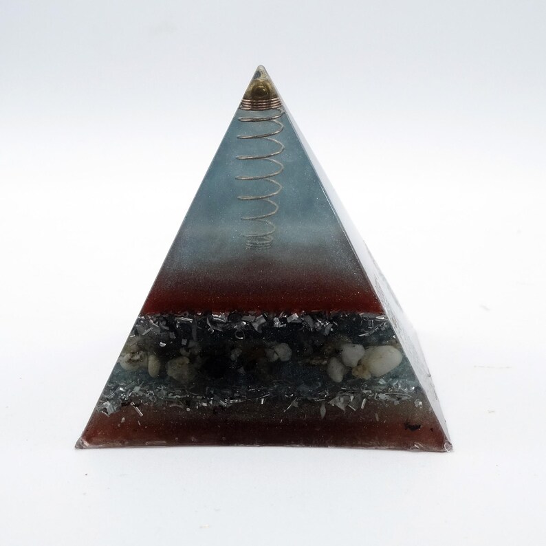 May include: A pyramid-shaped decorative object with a layered design. The top section is a gradient of blue and clear resin, followed by a red-brown layer, and a base with stones and dark inclusions. A coiled wire sits at the apex.
