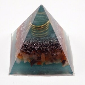 May include: An orgone energy pyramid, constructed from clear resin. The pyramid features layers of blue, brown, and black materials. A set of gold-coloured rings are embedded near the apex.