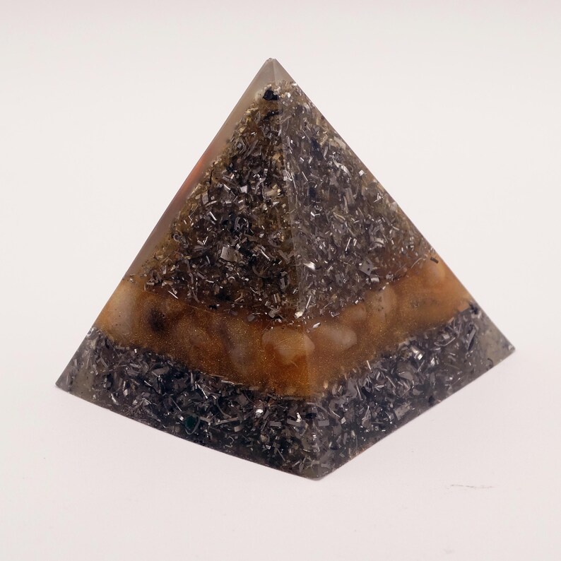 May include: A pyramid-shaped decorative object with a layered design. The top and bottom sections are filled with dark, metallic-looking fragments, while the middle section has a warm, amber-colored appearance. The pyramid is set against a plain white background.