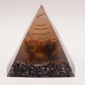 May include: A transparent pyramid-shaped object with a copper coil at the top and embedded stones. The base is filled with dark, shiny fragments. The pyramid is a decorative item, possibly for spiritual or aesthetic purposes.
