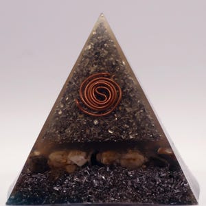 May include: A pyramid-shaped decorative object with a copper coil at the center. The pyramid is filled with dark, granular material and small stones. The pyramid is encased in a clear, translucent material.