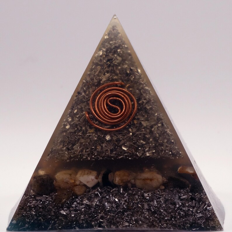 May include: A pyramid-shaped decorative object with a copper coil at the center. The pyramid is filled with dark, granular material and small stones. The pyramid is encased in a clear, translucent material.