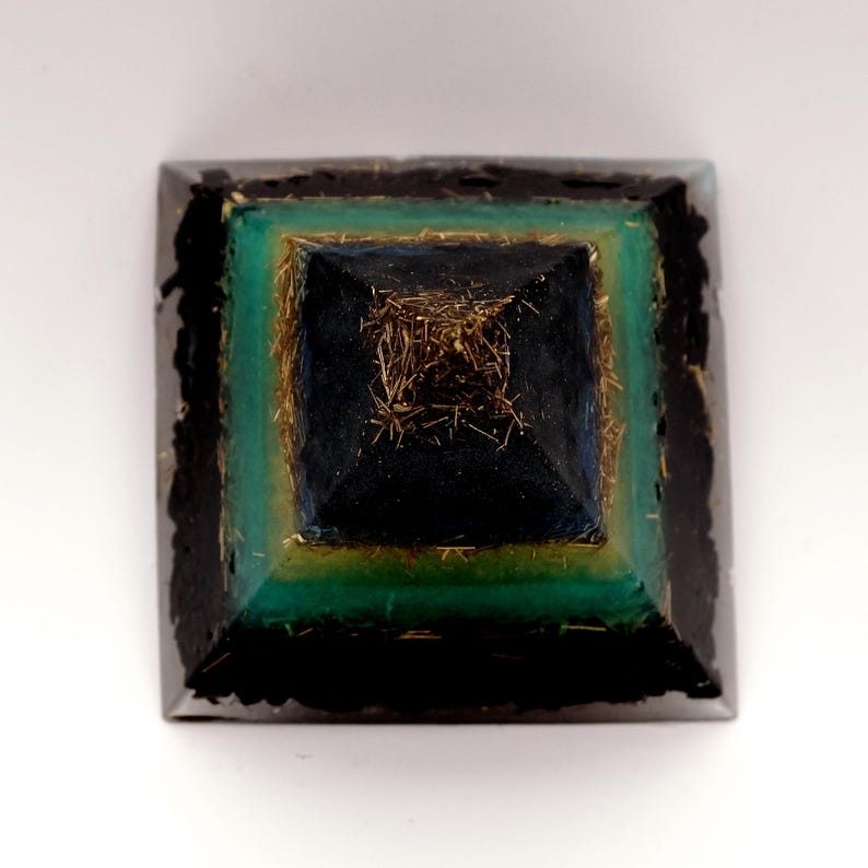 May include: A square, layered pyramid with a black base and a dark teal, gold, and black top. The pyramid has a textured appearance, with gold-colored strands visible in the center. The pyramid is set against a white background.