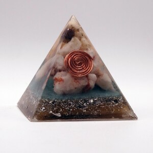 May include: A pyramid-shaped resin art piece with a copper coil at the center. The pyramid contains white and red stones, a teal layer, and a dark base with silver flecks. The pyramid is on a white background.