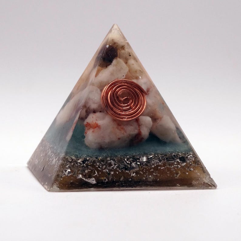 May include: A pyramid-shaped resin art piece with a copper coil at the center. The pyramid contains white and red stones, a teal layer, and a dark base with silver flecks. The pyramid is on a white background.