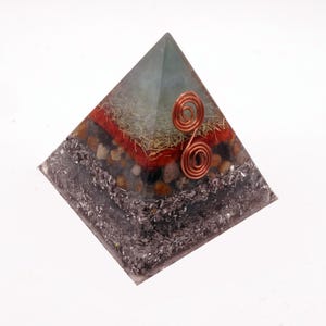 May include: A layered pyramid-shaped object with a copper wire spiral on the front. The pyramid has a clear, light green top, a red layer, and a dark gray base. The layers include small stones and metallic flakes.