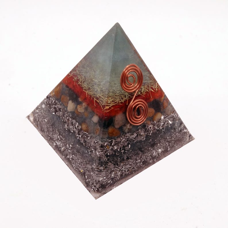 May include: A layered pyramid-shaped object with a copper wire spiral on the front. The pyramid has a clear, light green top, a red layer, and a dark gray base. The layers include small stones and metallic flakes.