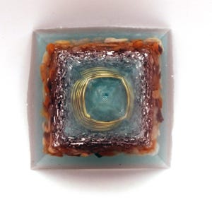 May include: A square orgone pyramid with a light blue resin base. It features layers of silver metal, brown and tan stones, and a central teal stone encased in gold wire. The pyramid is a decorative item, likely for display.