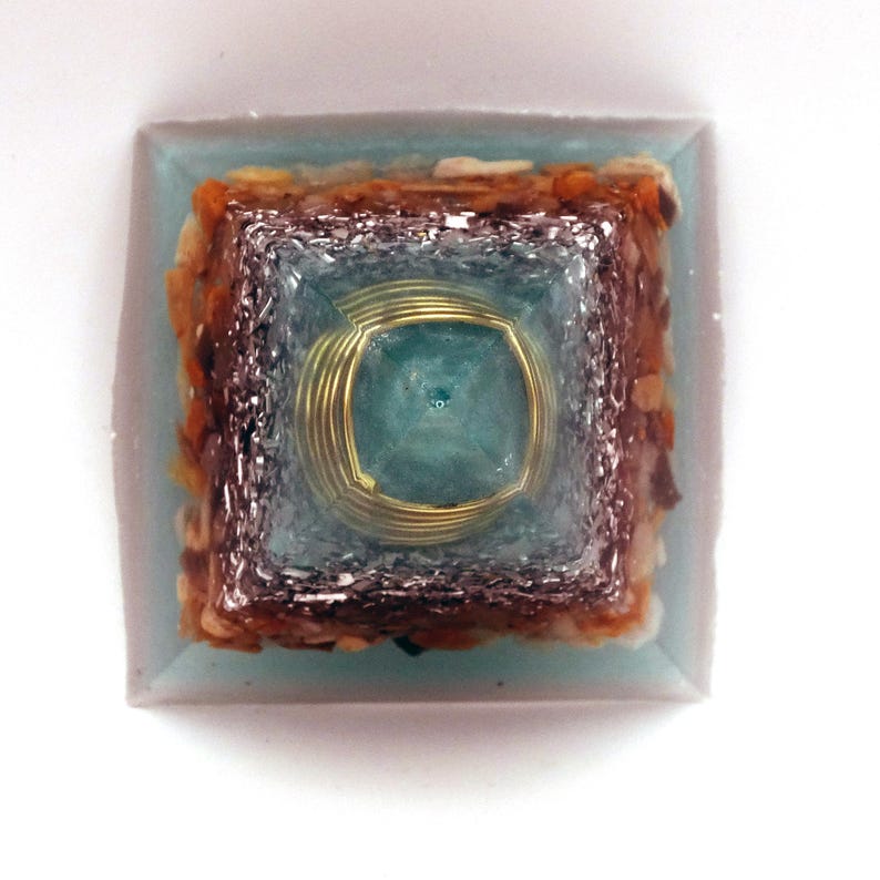 May include: A square orgone pyramid with a light blue resin base. It features layers of silver metal, brown and tan stones, and a central teal stone encased in gold wire. The pyramid is a decorative item, likely for display.