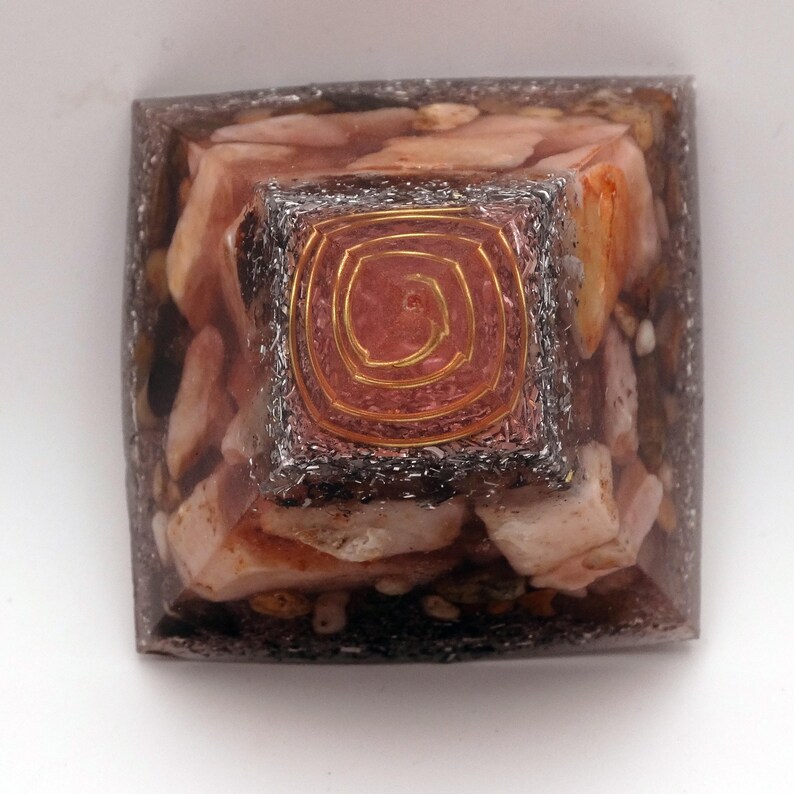 May include: A square orgone pyramid with a copper spiral on top. The pyramid is filled with pink and white stones and encased in a clear resin. The copper spiral is centered on a smaller, square, metallic-looking base.