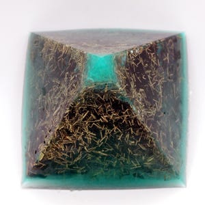 May include: A square orgone pyramid with a teal-colored resin exterior. The interior features a dark brown material with gold-colored, needle-like inclusions. The pyramid has a square base and a pointed top.