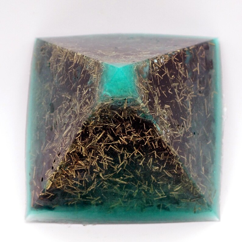 May include: A square orgone pyramid with a teal-colored resin exterior. The interior features a dark brown material with gold-colored, needle-like inclusions. The pyramid has a square base and a pointed top.