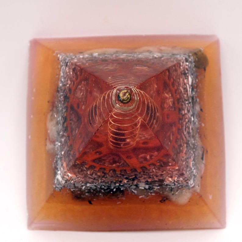 May include: A square-based pyramid with a copper-colored interior and a clear, amber-colored resin exterior. The pyramid's apex features a copper coil and a small, gold-colored element. The base is square, and the overall design is geometric.