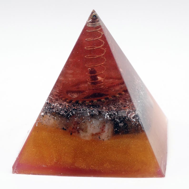 May include: A pyramid-shaped orgone energy generator. The pyramid is made of a translucent orange resin with a copper coil at the top. The base has layers of orange, black, and white materials.