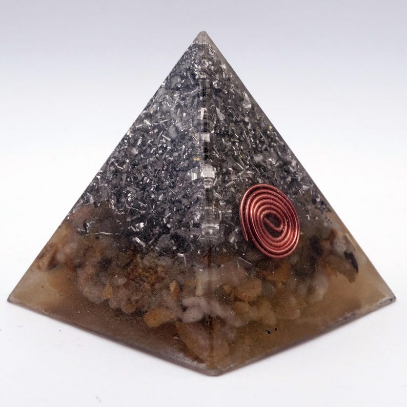 May include: A pyramid-shaped orgone energy generator. The top half is filled with dark, metallic-looking material, while the bottom half has a tan, stone-like appearance. A copper coil is embedded on the side. The pyramid is on a white background.