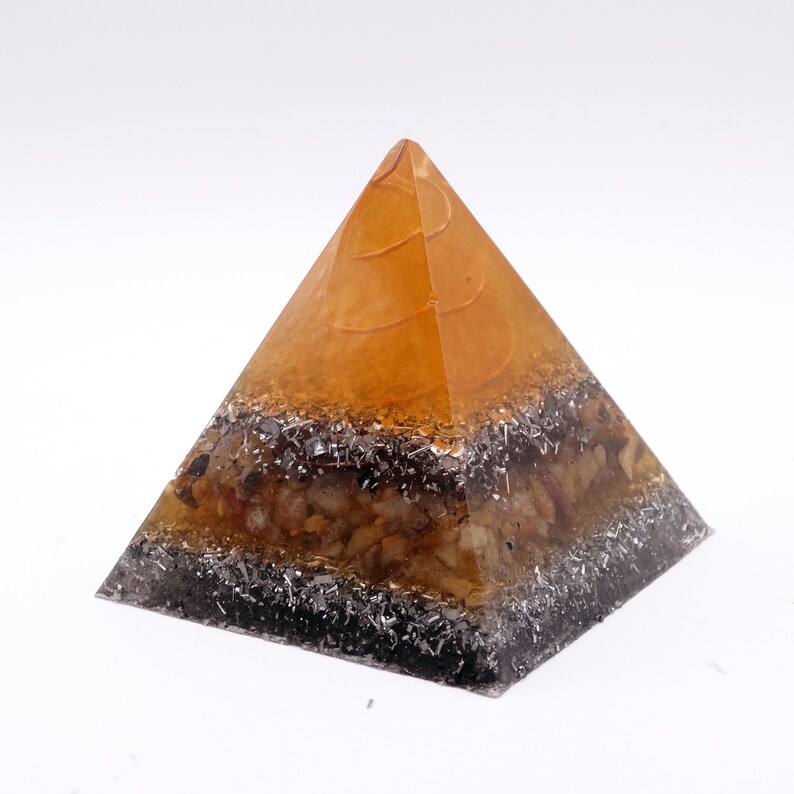 May include: A pyramid-shaped decorative object with a layered design. The top is amber-colored, transitioning to a layer of dark gray and silver, and a base of mixed-color stones. The pyramid is set against a white background.