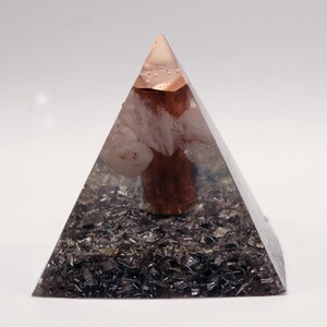 Op de afbeelding: Een heldere piramide met een koperkleurige top en een donkere basis. Binnenin bevinden zich lichtgekleurde stenen en een centraal, cilindrisch, roodbruin element. De piramide staat tegen een witte achtergrond.