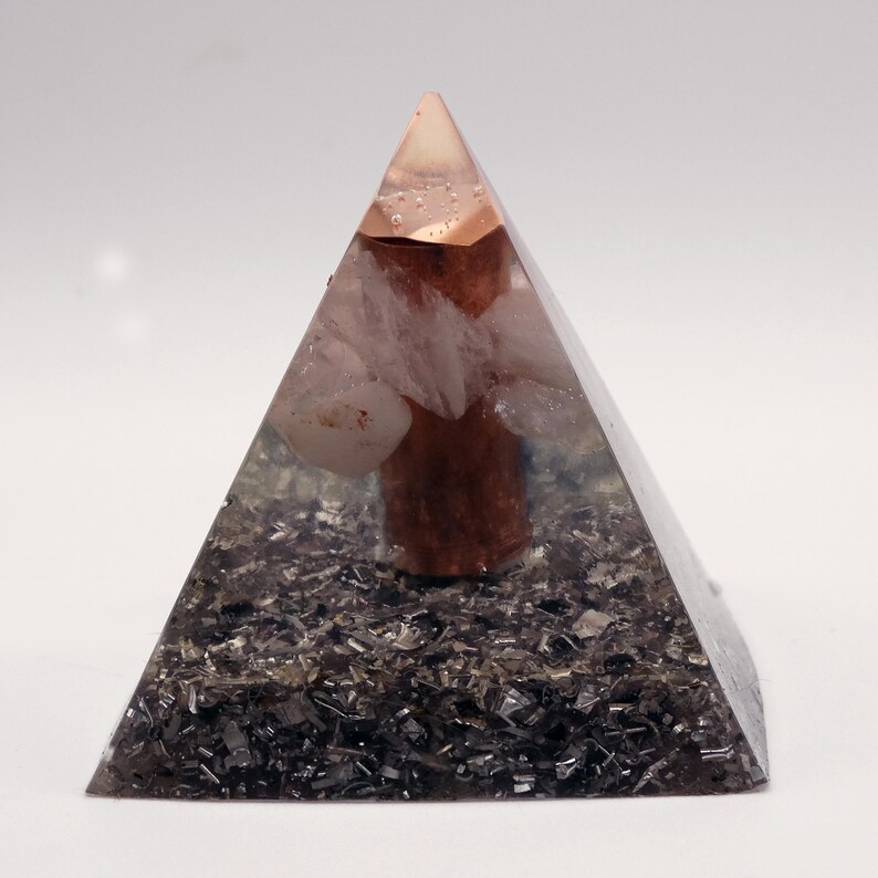Op de afbeelding: Een heldere piramide met een koperkleurige top en een donkere basis. Binnenin bevinden zich lichtgekleurde stenen en een centraal, cilindrisch, roodbruin element. De piramide staat tegen een witte achtergrond.