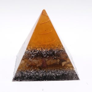 May include: A clear pyramid-shaped resin art piece with an amber-colored top and a layered interior. The layers include silver-colored metal flakes, dark brown and beige stones, and a dark base. The pyramid is displayed against a white background.