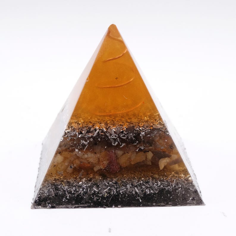 May include: A clear pyramid-shaped resin art piece with an amber-colored top and a layered interior. The layers include silver-colored metal flakes, dark brown and beige stones, and a dark base. The pyramid is displayed against a white background.