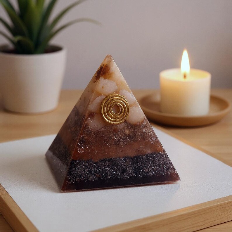 May include: A layered pyramid-shaped orgone energy generator with a gold spiral accent. The pyramid features alternating bands of brown, black, and cream-colored materials. A lit candle and a small potted plant are in the background.
