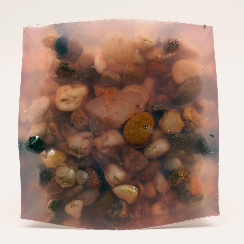 May include: A clear, square-shaped resin block filled with a variety of small, colorful pebbles. The pebbles range in color from white and tan to brown and green. The resin has a soft, pinkish hue, giving the block a unique appearance.