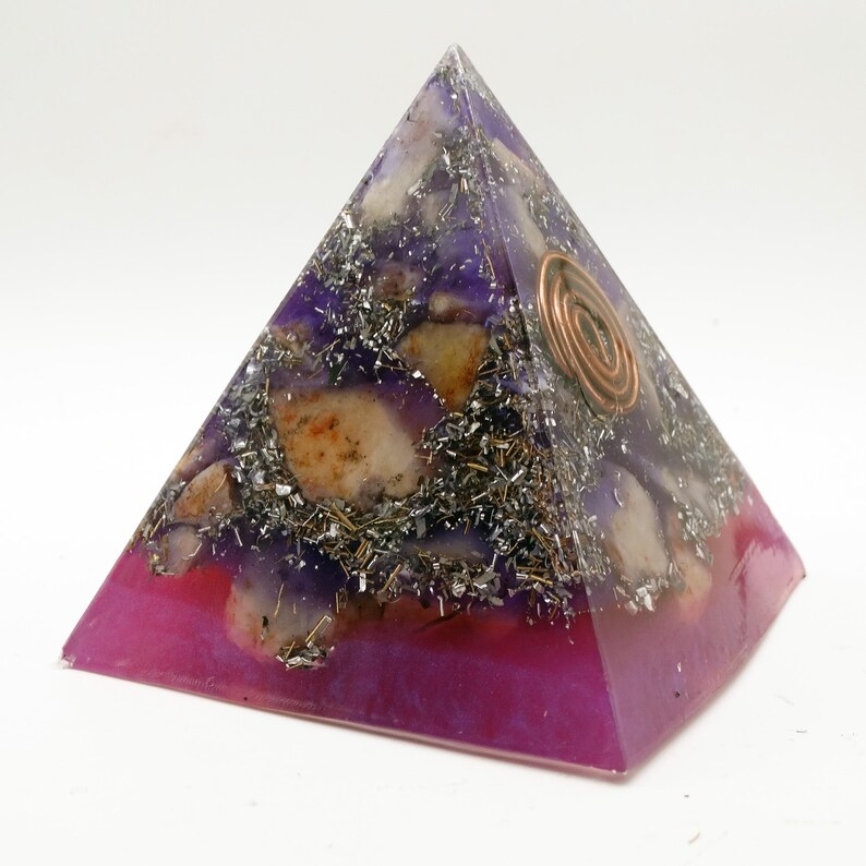 May include: A pyramid-shaped orgone energy generator. The base is a deep magenta, transitioning to purple with embedded stones and metal shavings. A copper coil is set on one side. The pyramid is a spiritual and decorative item.