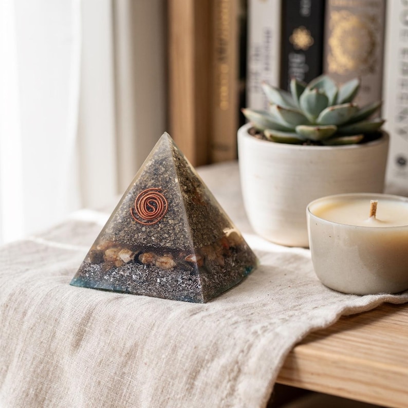 May include: A clear pyramid-shaped object with a copper coil embedded in the top. The pyramid is filled with small stones and a dark, granular material. It sits on a beige linen cloth, next to a small succulent plant in a white pot and a lit candle.