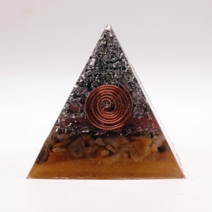 May include: A pyramid-shaped orgone energy generator. The pyramid features a copper coil at the center, encased in a clear resin with layers of brown, orange, and gray materials. The pyramid is designed to be a decorative and spiritual item.