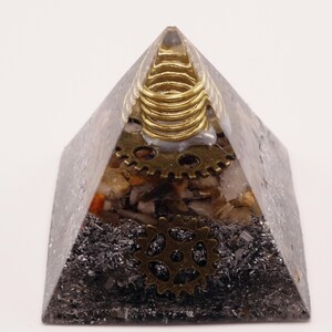 Puede incluir: Un objeto decorativo en forma de pir&aacute;mide con est&eacute;tica steampunk. La pir&aacute;mide est&aacute; hecha de un material transparente y gris con engranajes, piedras y detalles met&aacute;licos incrustados. Una bobina dorada se encuentra en la parte superior y se ven dos engranajes de lat&oacute;n.