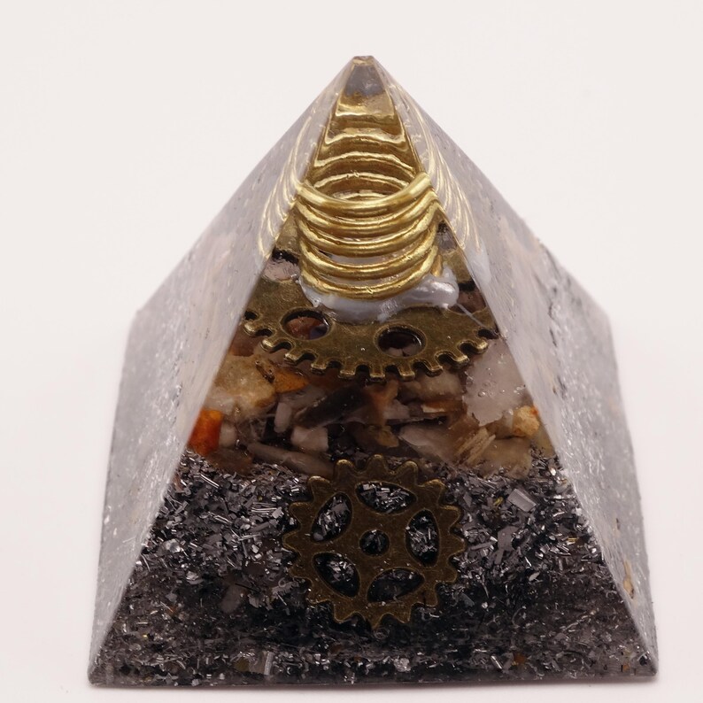 Puede incluir: Un objeto decorativo en forma de pir&aacute;mide con est&eacute;tica steampunk. La pir&aacute;mide est&aacute; hecha de un material transparente y gris con engranajes, piedras y detalles met&aacute;licos incrustados. Una bobina dorada se encuentra en la parte superior y se ven dos engranajes de lat&oacute;n.