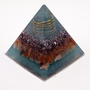 May include: A teal resin pyramid sculpture. It has a gold-coloured coil at the apex, with a mix of brown, beige, and silver-coloured stones and metal fragments embedded within the resin. The base is square.
