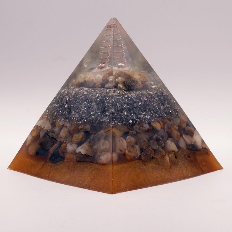 Puede incluir: Una pir&aacute;mide de resina transparente con capas de piedras, material gris oscuro y una bobina de cobre en el &aacute;pice. La base tiene un tono &aacute;mbar c&aacute;lido. Un art&iacute;culo de decoraci&oacute;n &uacute;nico.