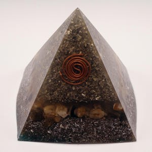 May include: A pyramid-shaped orgone device with a copper coil at the top. The pyramid is filled with a mix of dark gray and clear resin, with small stones and metallic flakes visible inside. The pyramid is on a white background.