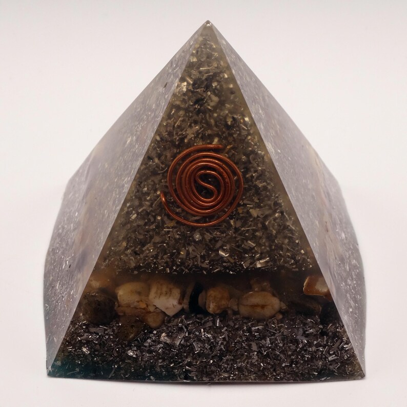May include: A pyramid-shaped orgone device with a copper coil at the top. The pyramid is filled with a mix of dark gray and clear resin, with small stones and metallic flakes visible inside. The pyramid is on a white background.