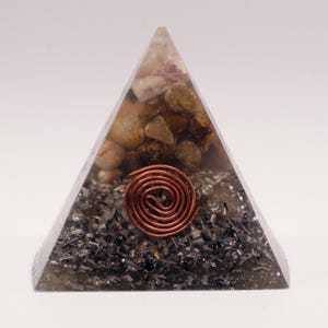 Puede incluir: Una escultura de resina transparente en forma de pir&aacute;mide con piedras incrustadas y una espiral de cobre. La pir&aacute;mide presenta una mezcla de piedras beige, marrones y grises, con una espiral de cobre en el centro.