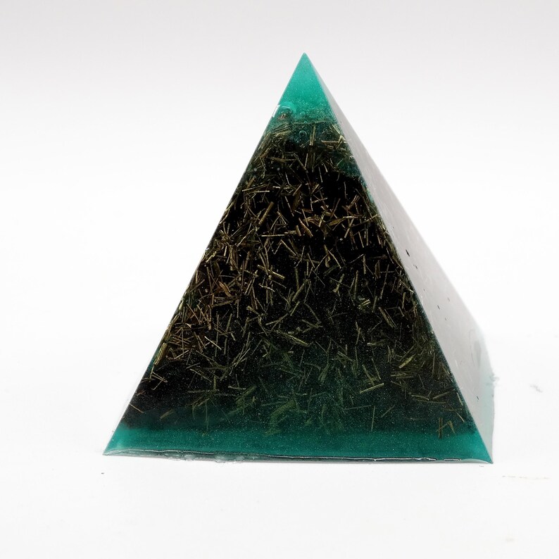 May include: A transparent pyramid with a teal-colored top and base. The interior is filled with dark, fibrous material, creating a striking contrast. The pyramid is set against a white background, highlighting its geometric form and unique composition.