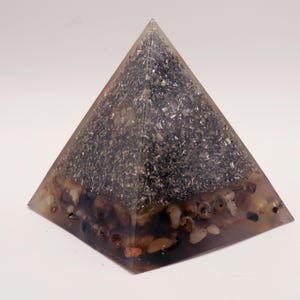 Puede incluir: Un objeto transparente en forma de pir&aacute;mide con un interior oscuro y granular y peque&ntilde;as piedras redondeadas en la base. La pir&aacute;mide tiene un exterior transparente, que muestra la composici&oacute;n interna. Las piedras son de varios tonos de marr&oacute;n y beige.