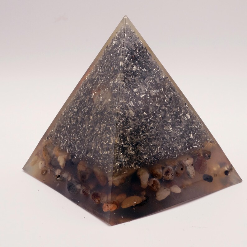 Puede incluir: Un objeto transparente en forma de pir&aacute;mide con un interior oscuro y granular y peque&ntilde;as piedras redondeadas en la base. La pir&aacute;mide tiene un exterior transparente, que muestra la composici&oacute;n interna. Las piedras son de varios tonos de marr&oacute;n y beige.
