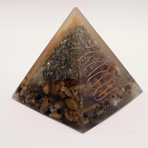 May include: A clear resin pyramid with embedded stones, metal flakes, and a copper coil. The pyramid has a layered appearance with various textures and colors, including beige, brown, and silver. The copper coil is a prominent feature.