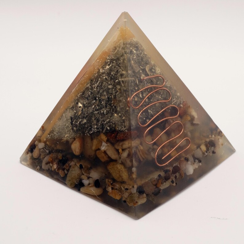 May include: A clear resin pyramid with embedded stones, metal flakes, and a copper coil. The pyramid has a layered appearance with various textures and colors, including beige, brown, and silver. The copper coil is a prominent feature.