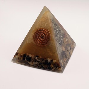 May include: A pyramid-shaped decorative object with a copper spiral embedded in the center. The pyramid is made of a clear resin with a tan and brown interior, and small stones at the base.